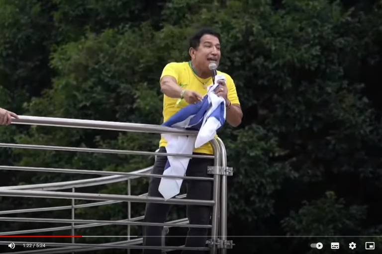 Magno Malta, de camiseta amarela e bandeira azul e branca na mão, está em pé, falando no microfone, apoiado em uma grade de metal. Ao fundo, copa de árvores