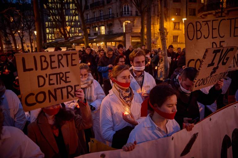Várias mulheres com mordaças na boca marcham em uma rua de Paris à noite. Em primeiro plano, uma delas ergue um cartaz de papelão onde se lê "liberdade de ser contra" em francês.