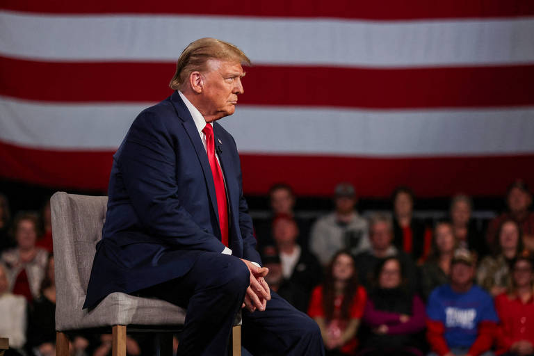 Trump é um homem branco de cabelos loiros, e está sentado, de terno, olhando para fora do quadro. Atrás dele há pessoas e uma enorme bandeira americana fora de foco.