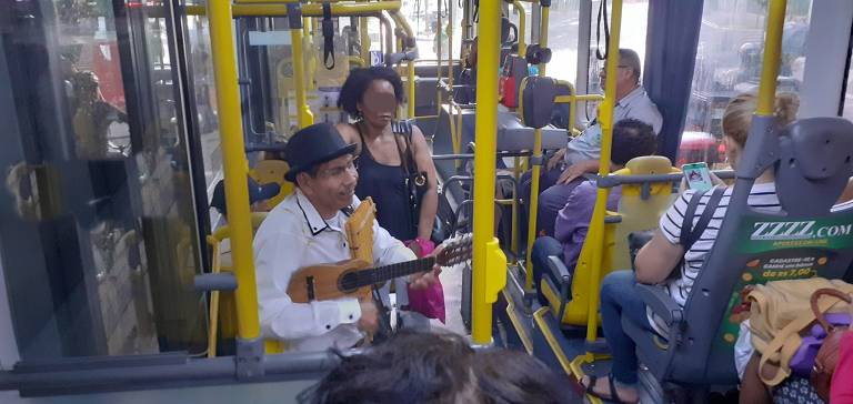 Em foto colorida, um músico aparece tocando charango, zampona e cantando dentro de um ônibus