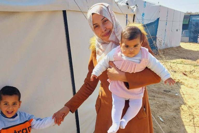 Foto mostra Taghreed, uma mulher árabe de pele clara que veste o hijab, o véu islâmico, sorrindo enquanto carrega no colo um bebê e dá as mãos para um menino de 5 anos de idade. Atrás dela é possível ver uma barraca do campo de refugiados onde está.