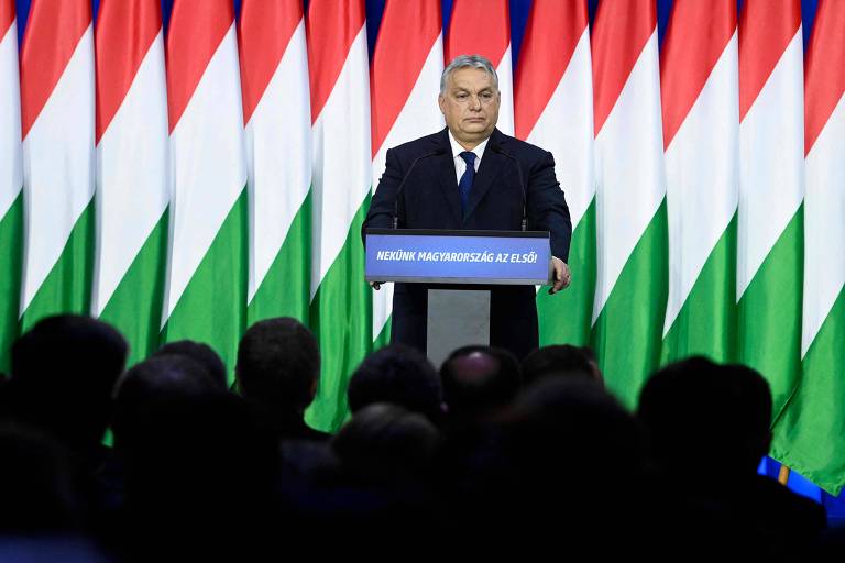 Homem de terno discursa em frente a púlpito num palco; atrás dele estão bandeiras da Hungrua