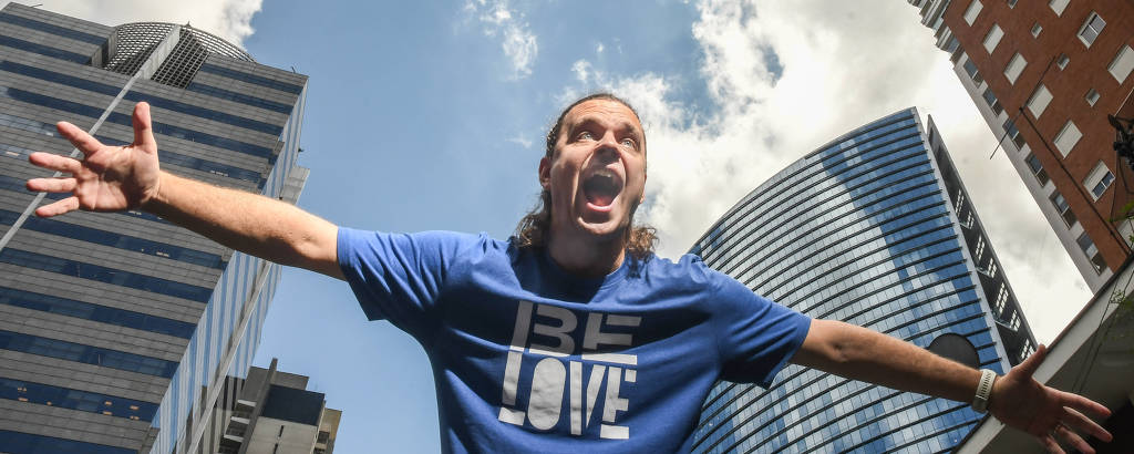 Homem branco com camiseta azul escrita Be Love posa com braços abertos com prédios ao fundo