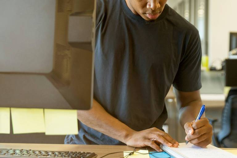 A imagem mostra um homem negro vestindo uma camiseta preta de manga curta. Ele escreve em em um caderno, que está em cima de uma mesa de madeira. Na sua frente, há um computador e um reclado