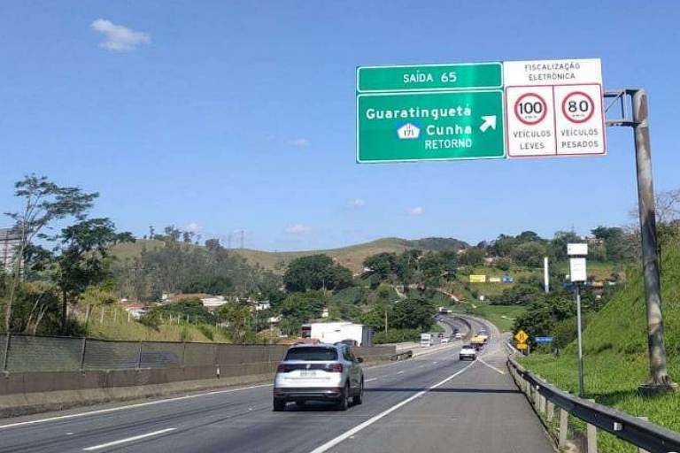 Imagem da estrada, com uma placa verde indicando a entrada de Guaratinguetá e um radar no acostamento à frente