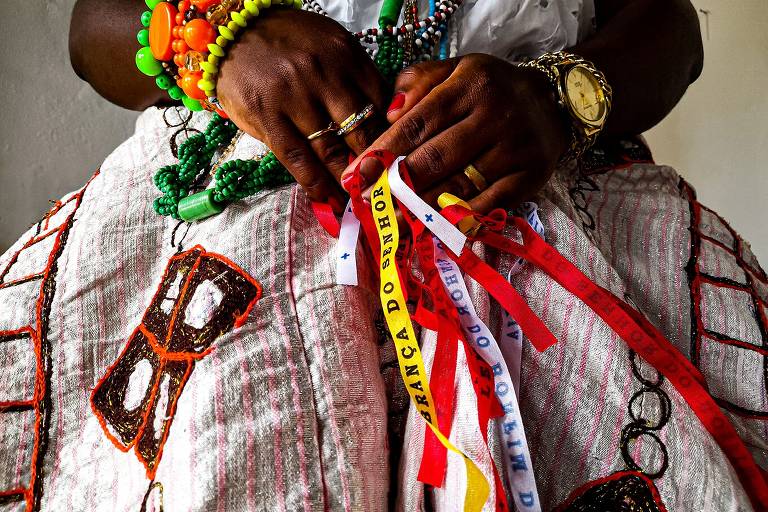 A foto mostra as mão de uma mulher segurando fitas coloridas do Senhor do Bonfim. Ela uso vestes tradicionais baianas e pulseiras coloridas no braço direito. No braço esquerdo, há um relógio dourado
