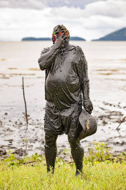 Um homem totalmente coberto de lama tampando o rosto. Em sua mão esquerda, segura um chapéu