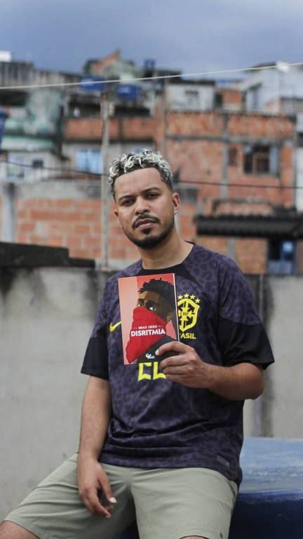 Homem negro de camisa preta e bermuda (Ronald Lincoln) posa diante de uma favela com livro de sua autoria, "Disritmia", na mão esquerda 