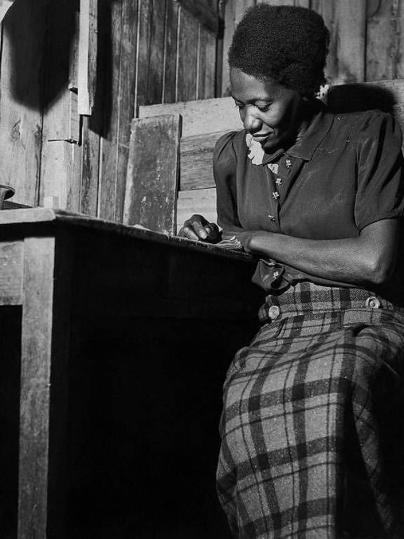 Carolina Maria de Jesus é registrada escrevendo sobre uma mesa de madeira. Ela é uma mulher negra que ocupa o canto da imagem. Está sentada, aparentemente absorta no que escreve.