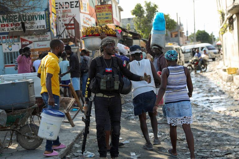 Foto mostra um homem negro armado com metralhadora e um colete onde se lê a palavra "polícia" gesticulando para que civis continuem a andar por uma rua