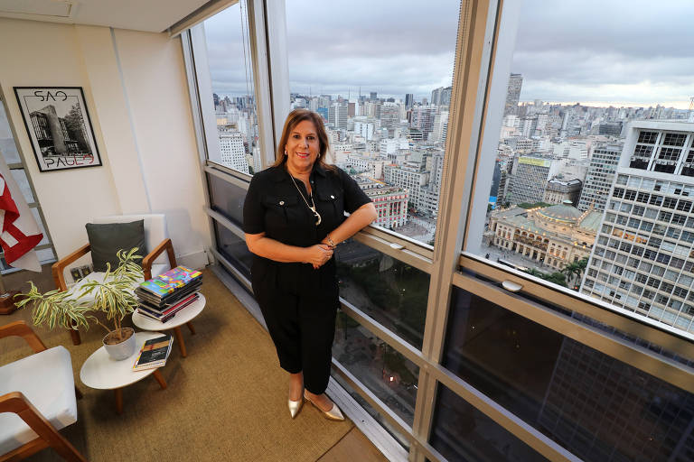 Mulher branca, vestida com calça e blusa pretas, cruza os braços à frente do corpo, que está apoiado a uma janela de vidro que dá um panorama da cidade de São Paulo