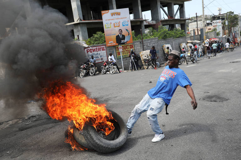 Um homem negro de jeans e camiseta chuta um pneu pegando fogo no meio de uma rua no Haiti.