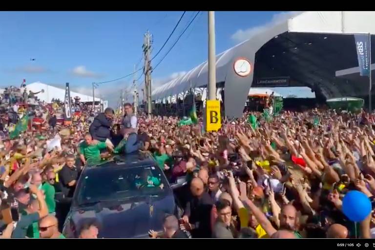 Multidão cerca carro em que está Jair Bolsonaro. Ele está de pé na caçamba. Ao fundo, céu azul