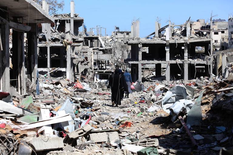Foto mostra prédios em ruínas, com escombros por toda parte, e duas figuras humanas caminhando por entre os destroços, ao longe