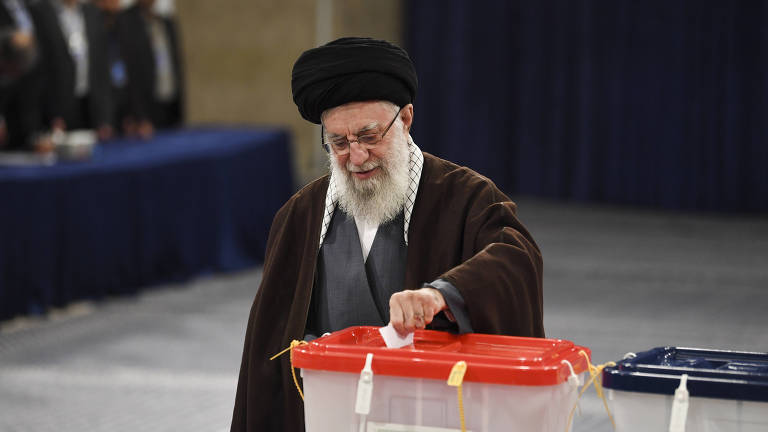 homem idoso de barba branca comprida, com vestes pretas típicas de líderes religioso do Irã, deposita uma cédula de papel em uma urna 
