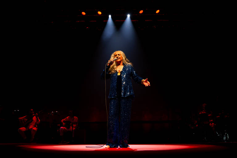 No Espaço Unimed, Maria Bethânia se apresenta sob um feixe de luz em um palco escuro, com a silhueta de músicos ao fundo. A cantora veste um conjunto de paetê azul escuro e está descalça no palco. Ela tem cabelo longo e grisalho