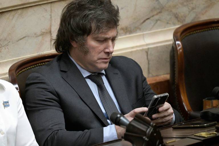 Homem de meia idade, branco, de cabelo castanho, vestindo terno escuro com gravata escura, olha para o celular, sentado
