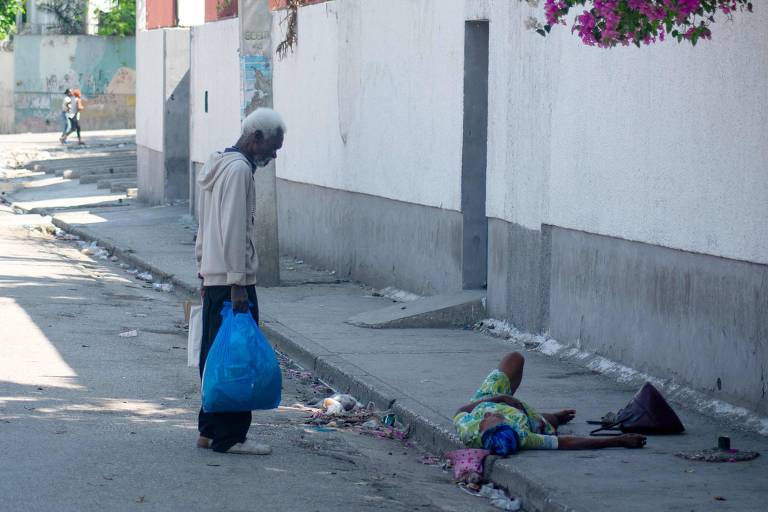 Homem negro, idoso, de camiseta cinza e carregando um saco azul, observa mulher negra, idosa, deitada em uma calçada