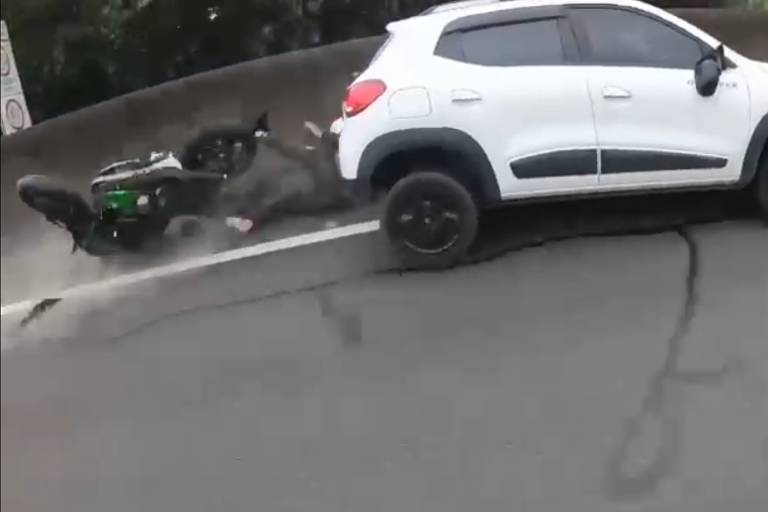 foto mostra carro trafegando em alta velocidade após atingir moto, é possível ver pernas do motoqueiro por trás do carro e moto caída