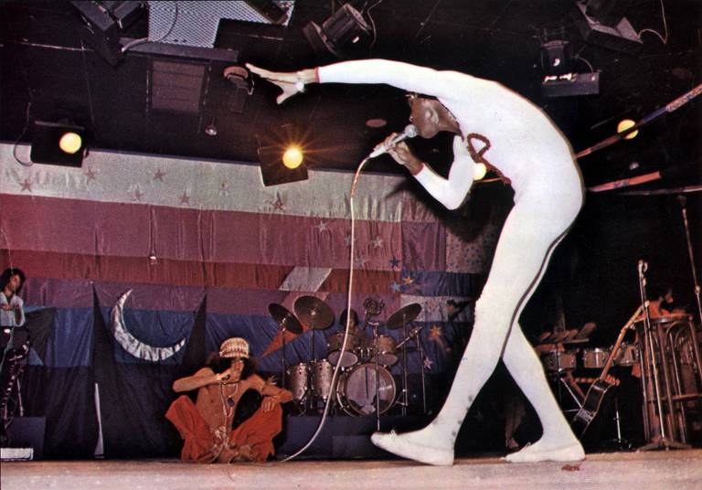 Em foto colorida, Gilberto Gil aparece cantando segurando um microfone em um palco onde ao fundo está sentado Caetano Veloso