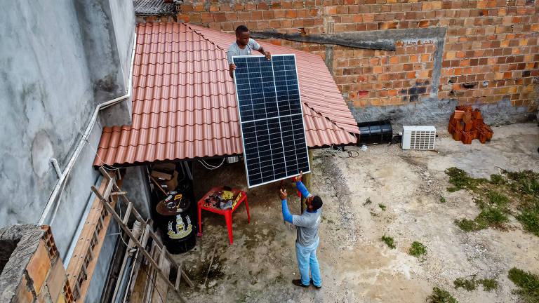 dois homens, um no telhado, seguram placa solar com parede sem reboco ao fundo