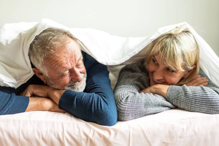 casal de homem e mulher idosos brancos deitados numa cama, sorrindo um para o outro