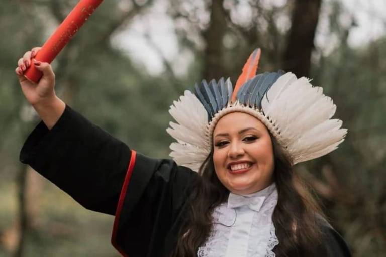 Mulher indígena levanta canudo de diploma. Ela usa uma beca preta e cocar com penas brancas, azul e vermelha na cabeça