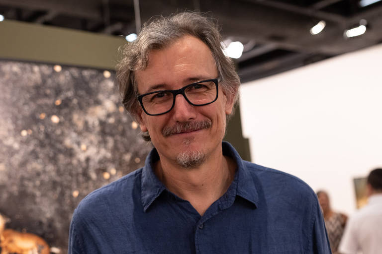 A imagem mostra um homem de meia-idade com cabelos grisalhos e óculos, vestindo uma camisa azul, sorrindo para a câmera. Ele está em uma galeria de arte, com uma obra de arte desfocada ao fundo e outras pessoas ao redor.
