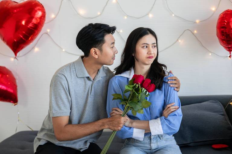 Fotografia mostra um casal de uma mulher e um homem asiáticos, sentados num sofá; ele se vira em direção a ela com um buquê de rosas vermelhas, mas ela olha para a direção oposta com expressão de contrariada e de braços cruzados