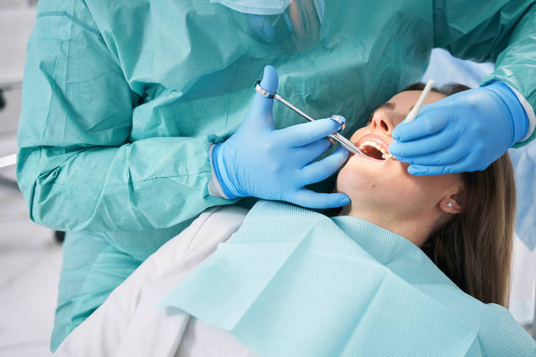 Fotografia de um dentista paramentado com avental e luvas verdes aplicando anestesia em uma paciente branca, deitada na cadeira