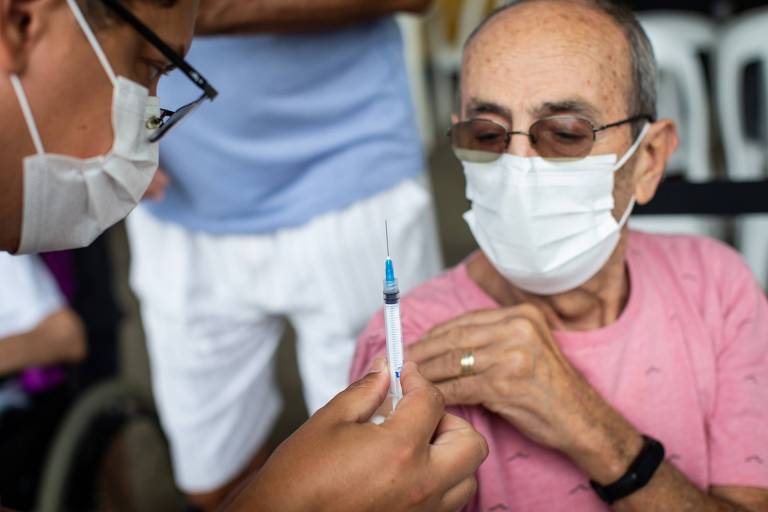 Idoso olha de camiseta rosa e máscara segura manga da camiseta enquanto observa seringa de vacina de gripe segurada por enfermeiro