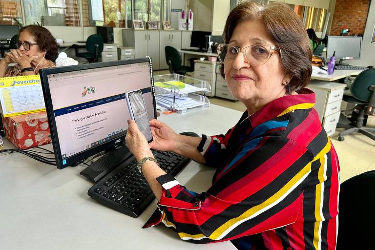 Mulher branca, de 67 anos, usa óculos enquanto segura um celular na frente de um computador numa repartição pública