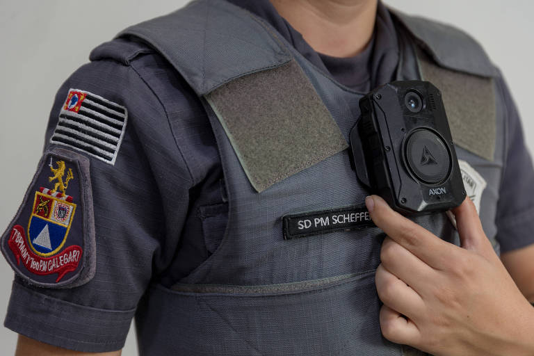 Imagem de um policial vestindo um uniforme cinza com um colete à prova de balas. No colete, há uma câmera corporal presa. No braço esquerdo do uniforme, há um emblema com uma bandeira e um brasão. O policial está segurando a câmera com a mão direita.

