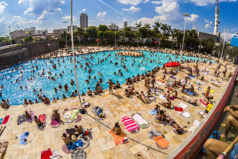 Dezenas de pessoas aproveitam grande piscina de cor azul, no Sesc Belenzeinho, em São Paulo. Há pessoas de roupas de banho dentro e ao redor da piscina. 