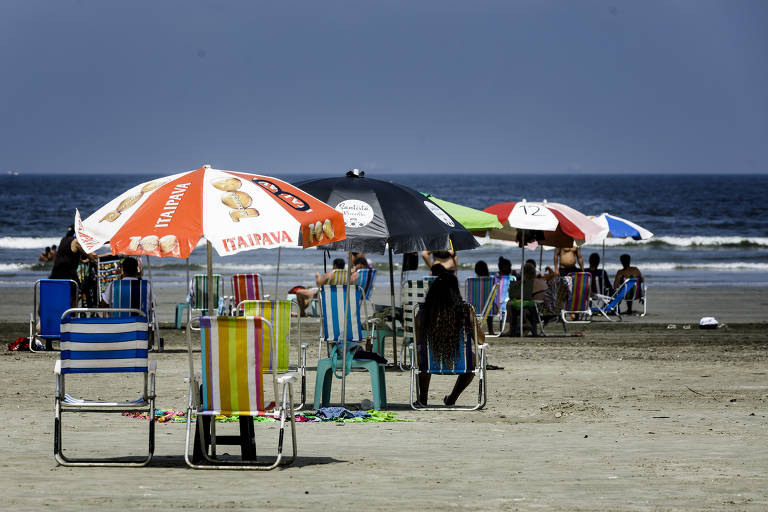 Cadeiras e guarda-sóis coloridos estão dispostos em uma praia; há poucas pessoas e o dia está bem iluminado