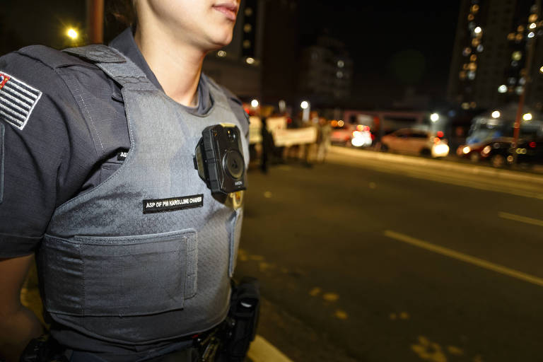 Foto de policial feminina em rua a noite na cidade de São Paulo. Vestida com sua farda e câmera ao centro dessa.