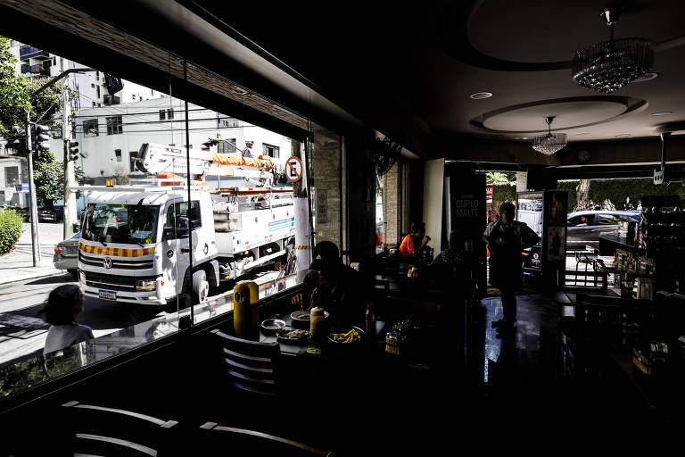 Lanchonete sem luz no bairro de Moema, em São Paulo, do lado de dentro, clientes estão sentados no escuro, enquanto do lado de fora, há o caminhão branco da companhia elétrica fazendo o reparo.