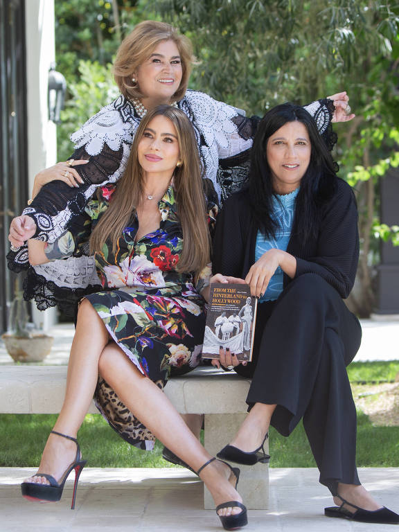 três mulheres posam para a foto; duas estão sentadas em um banco e uma terceira está atrás 