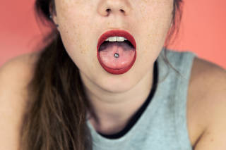 Close up portrait of young woman with freckles sticking out pierced tongue, showing her tongue piercing with pink background