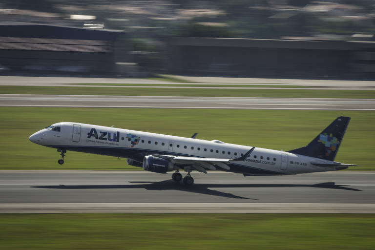 Um avião da companhia aérea Azul está decolando em uma pista de aeroporto. O avião é de fuselagem branca com detalhes em azul e verde. O fundo mostra uma área verde e uma pista de decolagem.