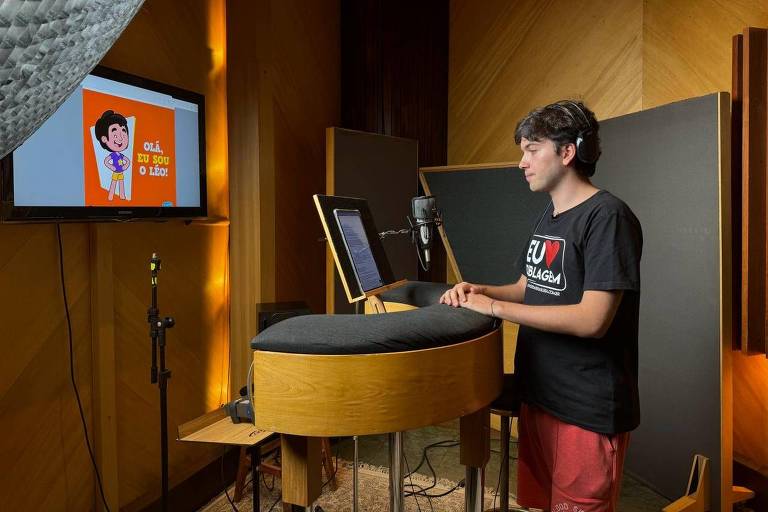 Homem jovem está de pé, com fones e olha para a tela do computador, de frente para um microfone