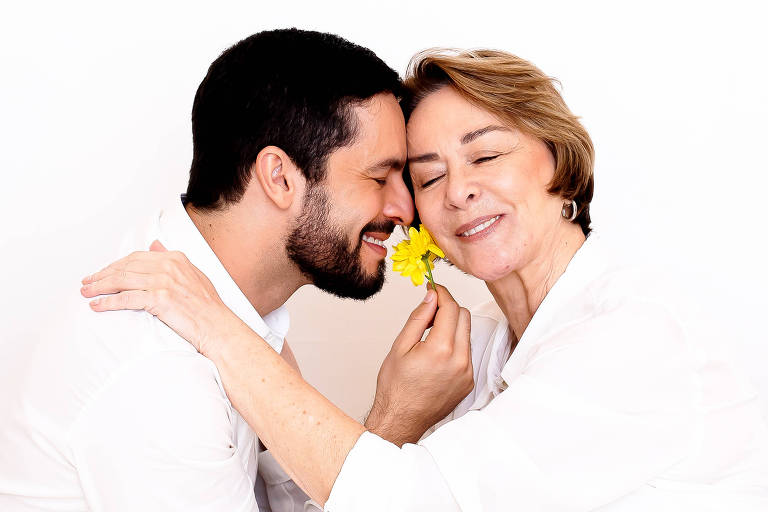 A imagem mostra um homem e uma mulher abraçados na frente de flores amarelas