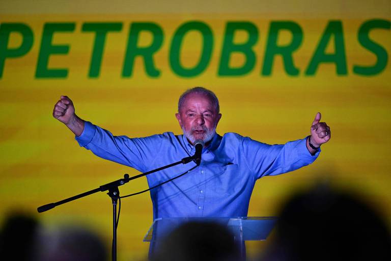 Um homem com expressão animada discursa energicamente diante de um fundo com o logotipo da Petrobras. Ele veste uma camisa azul e gesticula com os braços abertos.
