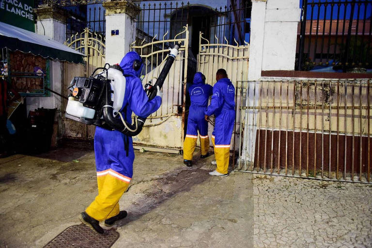 três pessoas com macacão azul e borrifador de veneno nas costas em portão de imóvel