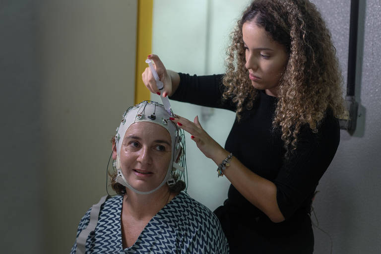 Eleitora de Lula veste uma touca de pano repleta de eletrodos para medição da atividade cerebral. Os eletrodos estão sendo preparados pela estudante Emanuelle Valadares, que manipula uma agulha próxima dos medidores