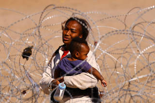 A migrant seeking asylum holds her baby as she stands near a razor wire fence deployed to prevent migrants from crossing into the United States, as seen from Ciudad Juarez