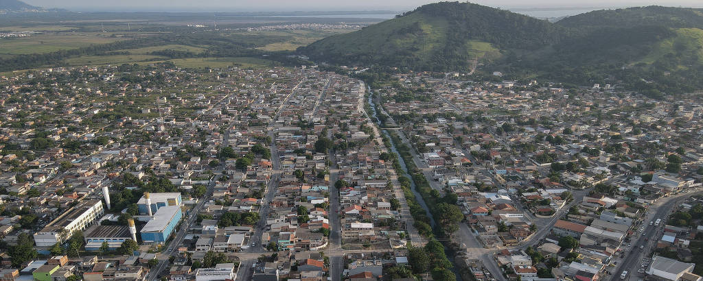 foto aérea mostra área densamente urbanizada, com muitas casas nas quadras, e, ao fundo da imagem, uma área verde vazia, ao longe, e dois morros com vegetação à direita, ao fundo 