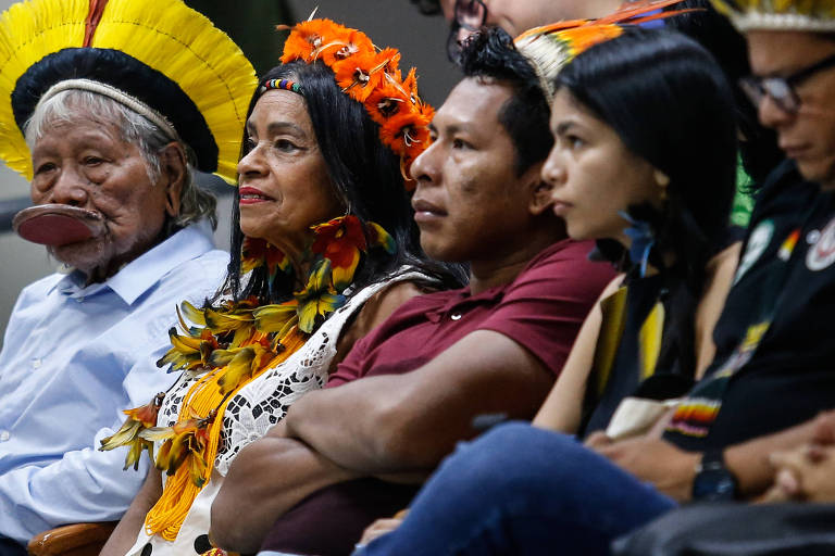 A imagem mostra um grupo de pessoas indígenas sentadas em um evento. À esquerda, um homem idoso com cabelo grisalho e um chapéu tradicional. Ao seu lado, uma mulher com cabelo longo e uma coroa de flores, vestindo roupas coloridas. Um homem jovem, com expressão séria, está ao centro, usando uma camiseta marrom. À direita, uma mulher com cabelo escuro e liso, vestindo uma blusa preta. Todos parecem estar atentos ao que acontece ao redor.