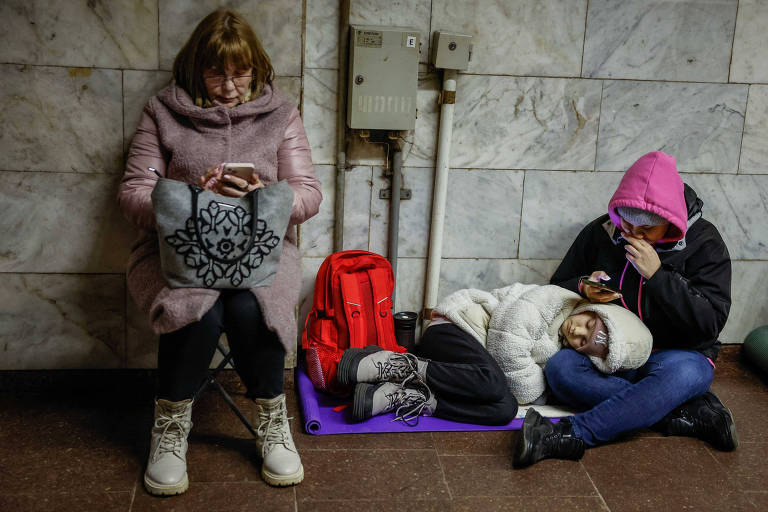 Escondida no metrô de Kiev para fugir de ataque russos, mulher checa o celular. A seu lado, criança dorme no colo de outra pessoa. Todos usam roupas de frio.