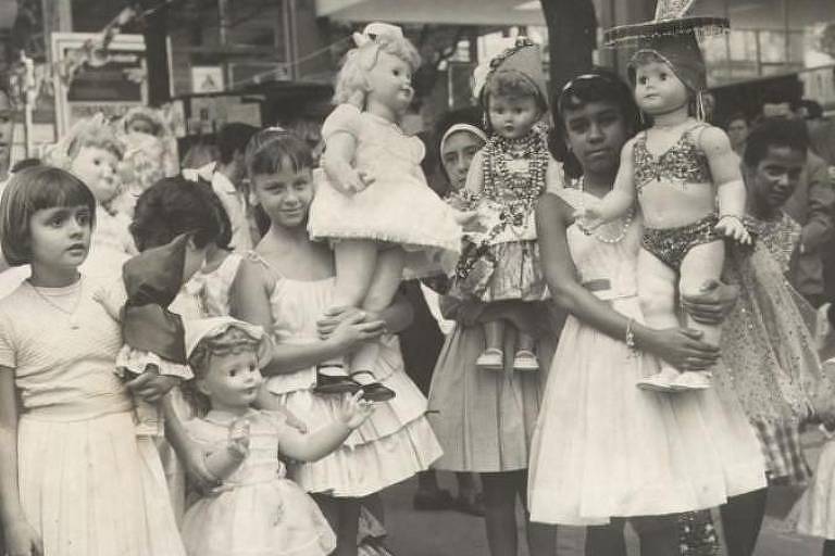 Foto em preto e branco mostra meninas brancas e negras de vestido segurando bonecas antigas